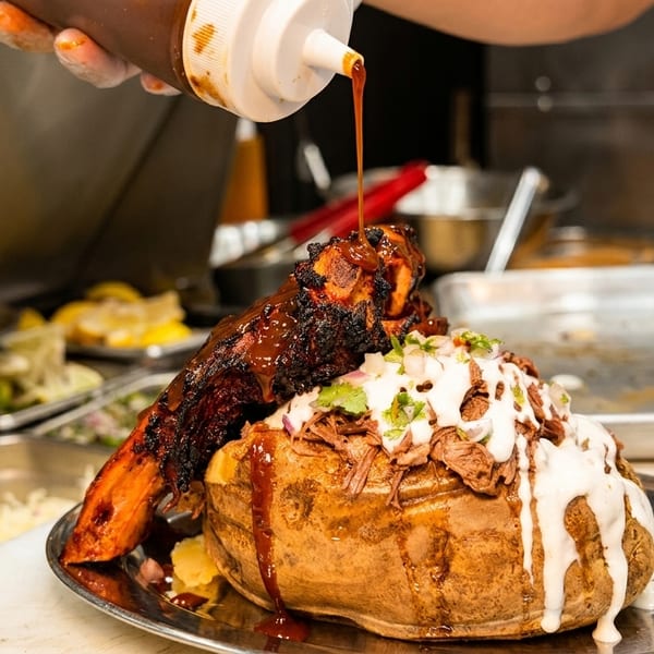 Chef pouring BBQ sauce over a loaded baked potato with ribs, topped with meat and white sauce.