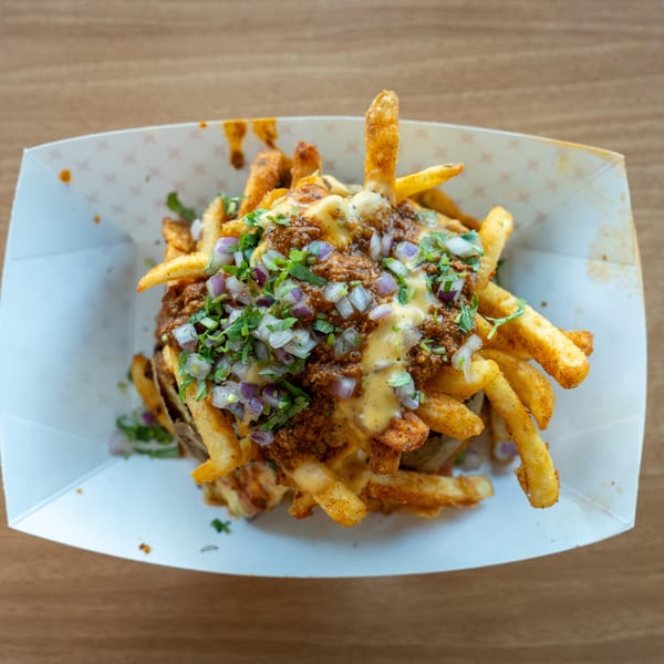 Chili cheese fries topped with diced red onions and cilantro in a white paper tray.