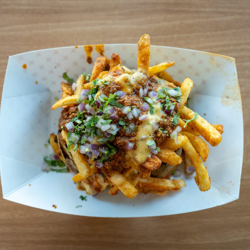 Chili cheese fries topped with diced red onions and cilantro in a white paper tray.