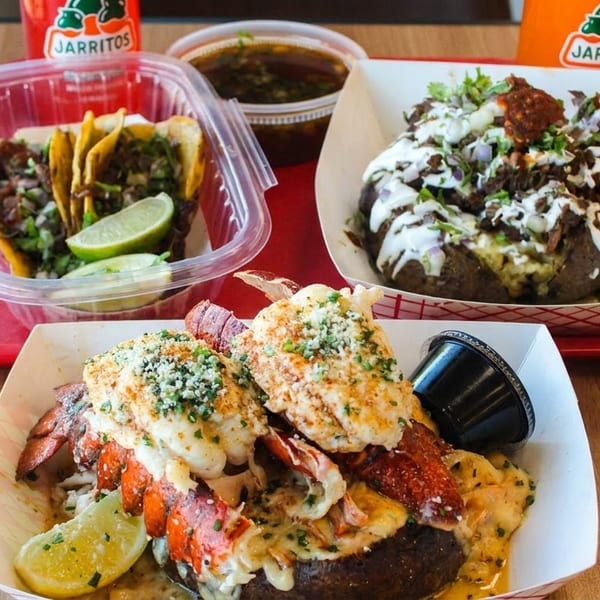 Tacos, loaded baked potatoes with lobster and meat, and Jarritos drinks on a wooden table.