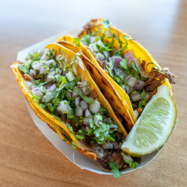 Three street tacos with meat, cilantro, and diced onions, served with a lime wedge.