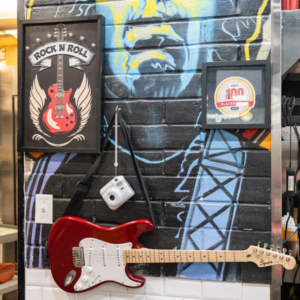 A red electric guitar hangs on a graffiti-covered brick wall with a "Rock N Roll" sign and Yelp award.