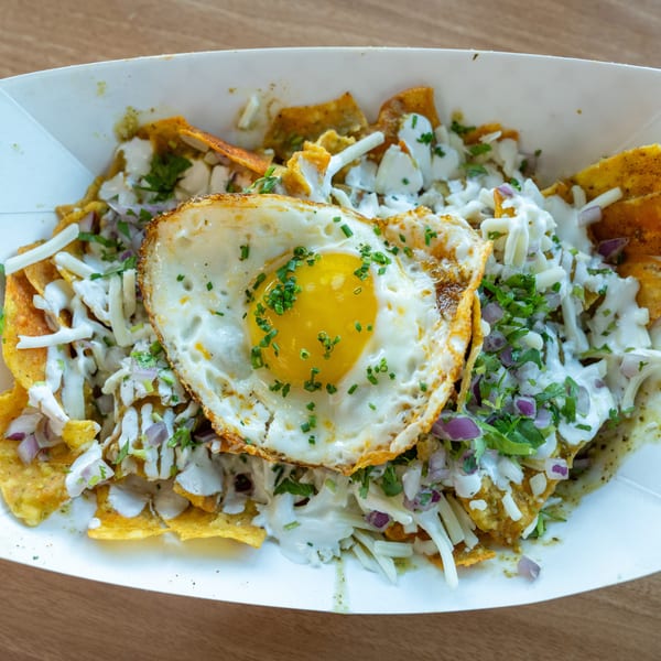 Chilaquiles with a fried egg, cheese, red onion, and cilantro in a white paper tray.
